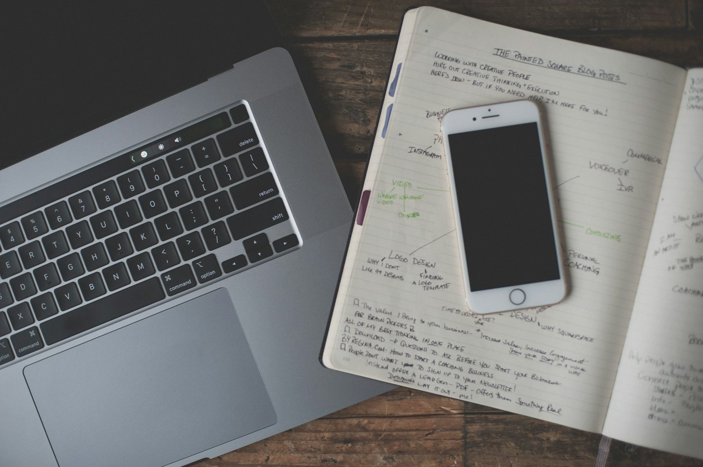 Laptop perak dan buku catatan dengan tulisan tangan di atas meja kayu.