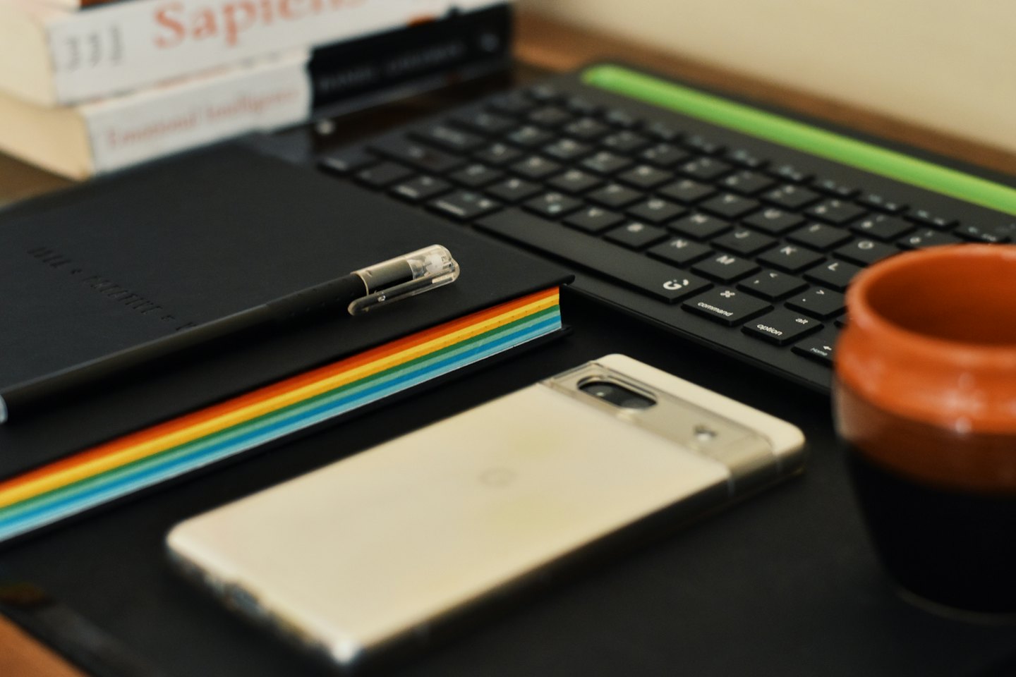 Sebuah buku catatan, pulpen, ponsel pintar, dan keyboard di atas meja.